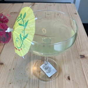May include: A clear glass margarita glass with a yellow and green paper umbrella. The glass is filled with a light green liquid.