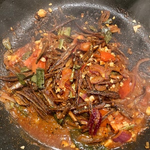 May include: A close-up of a black skillet filled with a mixture of fried fish, onions, tomatoes, and green herbs. The food is cooked in a brown sauce and appears to be steaming.