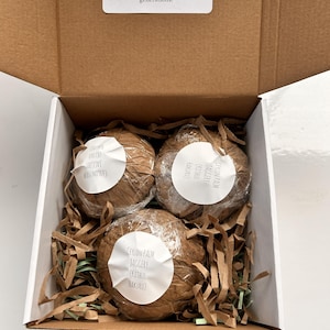 Puede incluir: Una caja de cartón abierta que contiene tres objetos redondos envueltos. Cada objeto está envuelto en papel marrón y tiene una etiqueta blanca. La caja también contiene papel triturado marrón y verde. Texto en una etiqueta: "A gift of pure Kithul jaggery..."