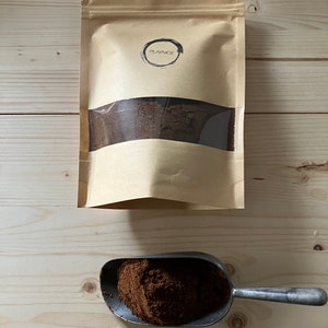 May include: A brown paper bag with a clear window, filled with dark brown powder, sits above a metal scoop filled with the same powder. The bag has the word "PLAYMOE" printed on it. The items are on a light wood surface.