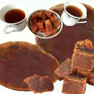 Puede incluir: Una vista aérea muestra dos tazas de té oscuro, un cuenco pequeño con cubos de azúcar moreno y varios trozos de azúcar moreno. El azúcar es de color marrón oscuro y las tazas de té tienen un diseño floral.