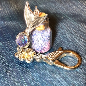 Könnte beinhalten: Ein silberner Meerjungfrauen-Schwanz-Anhänger, der an einer kleinen Glasflasche mit schimmerndem Glitzer befestigt ist. Die Flasche hat einen Korkverschluss und ist an einem silbernen Karabinerhaken befestigt.