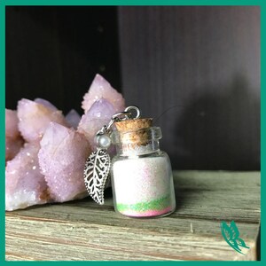 Puede incluir: Una pequeña botella de vidrio llena de purpurina blanca con acentos rosados y verdes. La botella tiene un tapón de corcho y está unida a un amuleto plateado con un diseño de hoja.