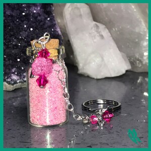 以下が含まれることがあります： ピンクのキラキラで満たされた小さなガラス瓶で、コルク栓で蓋がされています。銀色のチェーンが瓶を銀色のキーリングに繋いでおり、キーリングにはピンクのビーズが付いています。