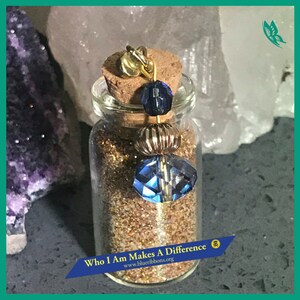 Peut inclure: Une petite bouteille en verre remplie de paillettes dorées et surmontée d'un bouchon. La bouteille a un charme bleu et or suspendu. La bouteille porte une étiquette qui dit "Who I Am Makes A Difference" et "www.blueribbons.org".