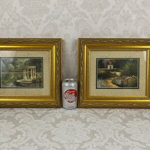 May include: Two framed pictures with gold frames. The pictures are of garden scenes. The picture on the left shows a gazebo in a garden with a pond. The picture on the right shows a garden path leading to a small cottage.