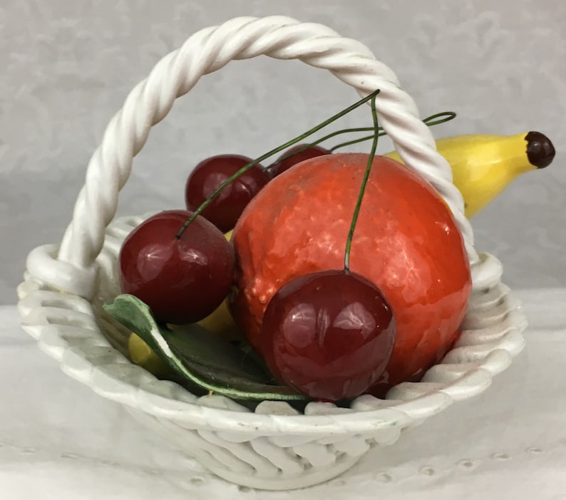Może przedstawiać: Biały ceramiczny koszyk z uchwytem w kształcie sznura wypełniony r&oacute;żnymi owocami, w tym czerwoną pomarańczą, wiśniami i ż&oacute;łtym bananem.