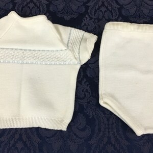 May include: A two-piece baby outfit in cream-colored knit fabric. The top has short sleeves and a textured panel across the chest with light blue accents. The matching bottoms are high-waisted shorts.