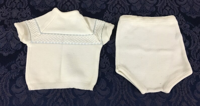 May include: A two-piece baby outfit in cream-colored knit fabric. The top has short sleeves and a textured panel across the chest with light blue accents. The matching bottoms are high-waisted shorts.
