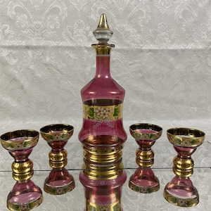 May include: A vintage pink glass decanter set with a stopper and four matching glasses. The set features gold accents and floral designs. The decanter and glasses are displayed on a mirrored surface.