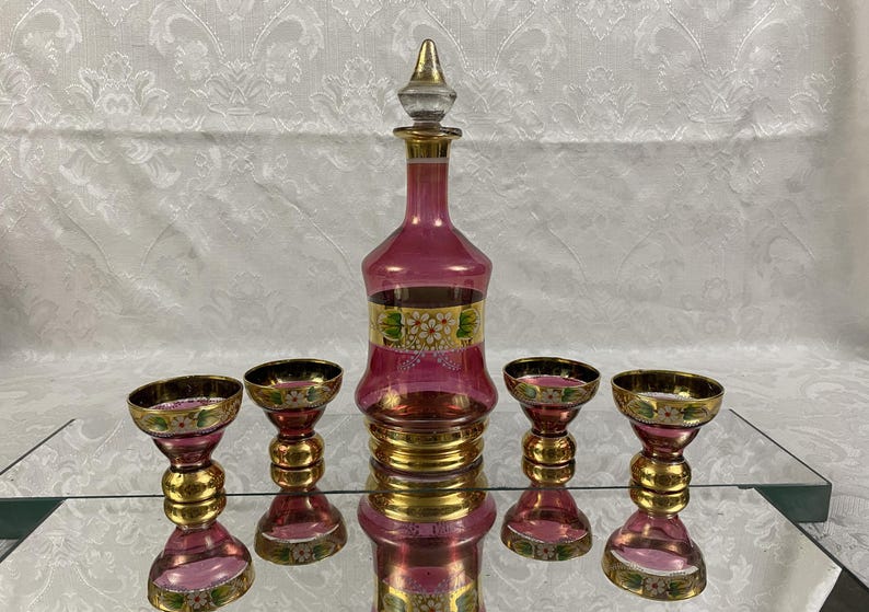 May include: A vintage pink glass decanter set with a stopper and four matching glasses. The set features gold accents and floral designs. The decanter and glasses are displayed on a mirrored surface.