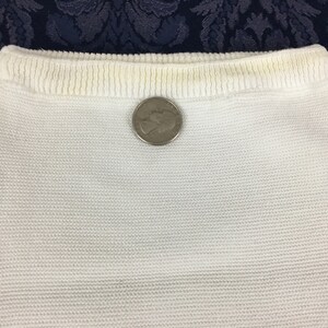 May include: Close-up of a white knitted fabric, possibly a garment or part of one. The fabric has a textured appearance. A silver quarter coin is placed on the fabric for size comparison.