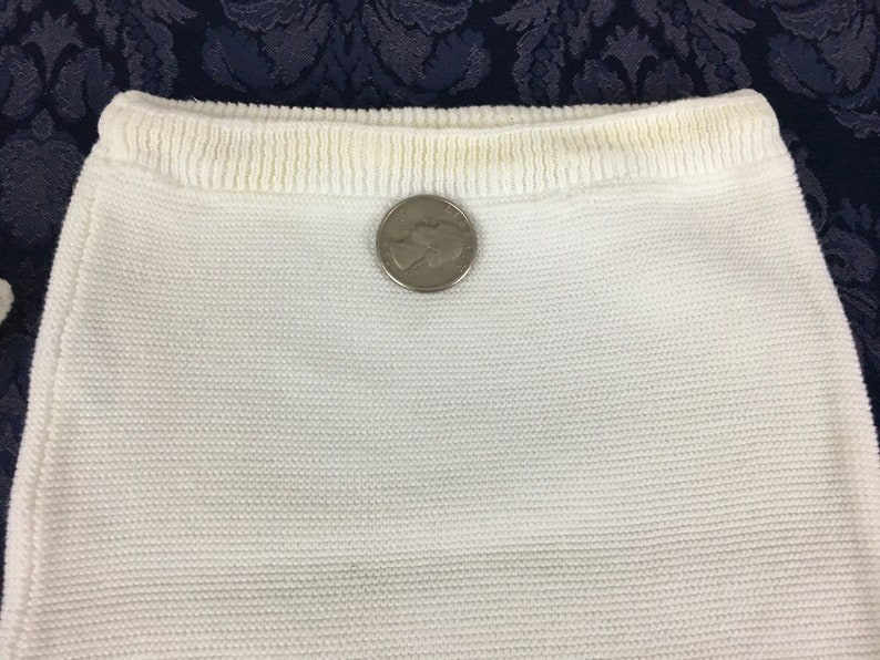 May include: Close-up of a white knitted fabric, possibly a garment or part of one. The fabric has a textured appearance. A silver quarter coin is placed on the fabric for size comparison.