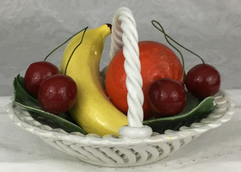 Może przedstawiać: Biały ceramiczny kosz na owoce z plecionym uchwytem. Kosz zawiera ż&oacute;łty banan, pomarańczowy pomarańczowy i cztery czerwone wiśnie.