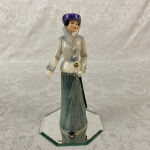 May include: A vintage porcelain figurine of a woman in a long skirt and jacket, holding a cane. The figurine wears a purple hat and is standing on a mirrored base. The jacket is white with blue accents and a gold button.