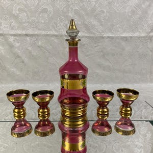 May include: A vintage pink glass decanter set with gold accents. The set includes a tall, ornate decanter with a stopper and four matching glasses. The decanter and glasses have a pink hue with gold rims and detailing.