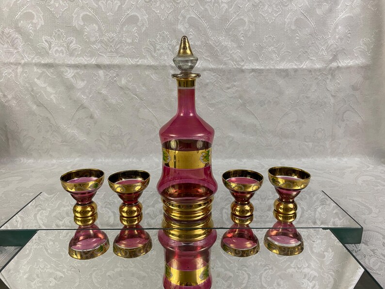May include: A vintage pink glass decanter set with gold accents. The set includes a tall, ornate decanter with a stopper and four matching glasses. The decanter and glasses have a pink hue with gold rims and detailing.