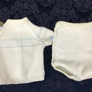 May include: A white knitted baby outfit, consisting of a short-sleeved top and matching shorts. The top features a textured design across the chest and light blue accents. The shorts have a ribbed waistband.