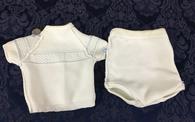 May include: A white knitted baby outfit, consisting of a short-sleeved top and matching shorts. The top features a textured design across the chest and light blue accents. The shorts have a ribbed waistband.