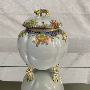 May include: A white porcelain jar with a lid, decorated with floral patterns in shades of orange, yellow, and blue. The jar has three gold-painted feet and a gold-painted handle on the lid.