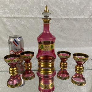 May include: A vintage pink glass decanter set with a matching stopper and four glasses. The set features gold accents and floral designs. A Diet Sam's can is next to the set.