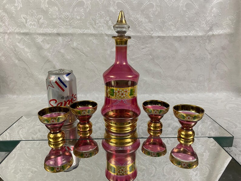 May include: A vintage pink glass decanter set with a matching stopper and four glasses. The set features gold accents and floral designs. A Diet Sam's can is next to the set.
