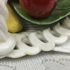 Może przedstawiać: Biały ceramiczny kosz na owoce z zakręconym uchwytem. Kosz jest ozdobiony ż&oacute;łtym bananem, czerwonym jabłkiem i zielonymi liśćmi. Kosz stoi na białym tiulowym obrusie.