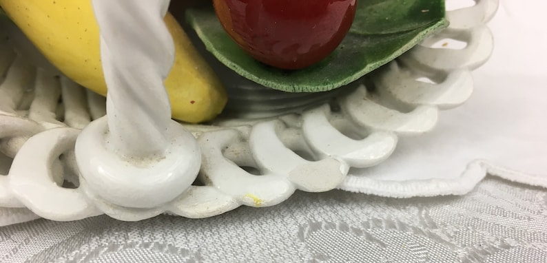Może przedstawiać: Biały ceramiczny kosz na owoce z zakręconym uchwytem. Kosz jest ozdobiony ż&oacute;łtym bananem, czerwonym jabłkiem i zielonymi liśćmi. Kosz stoi na białym tiulowym obrusie.