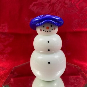 May include: A whimsical snowman figurine crafted from white glass, featuring a vibrant blue hat. The snowman has a carrot nose, rosy cheeks, and black dot eyes and mouth. The figurine is displayed on a reflective surface against a red backdrop.