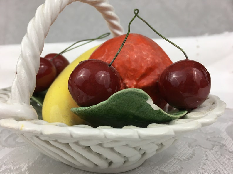 Może przedstawiać: Biały ceramiczny kosz na owoce z plecionym uchwytem. Kosz zawiera ż&oacute;łty banan, pomarańczowy pomarańczowy i trzy czerwone wiśnie.