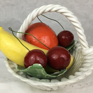 Może przedstawiać: Biały ceramiczny koszyk z plecionym uchwytem zawiera ceramiczną kompozycję owoc&oacute;w. Kompozycja zawiera ż&oacute;łty banan, czerwone jabłko i trzy czerwone wiśnie.