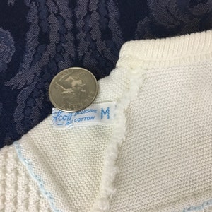 May include: Close-up of a white knit garment with a light blue trim. A quarter coin rests on the garment near a tag that reads "Accol Exclusive All Cotton M". The garment is on a dark blue patterned surface.