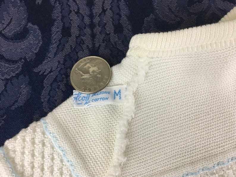 May include: Close-up of a white knit garment with a light blue trim. A quarter coin rests on the garment near a tag that reads "Accol Exclusive All Cotton M". The garment is on a dark blue patterned surface.