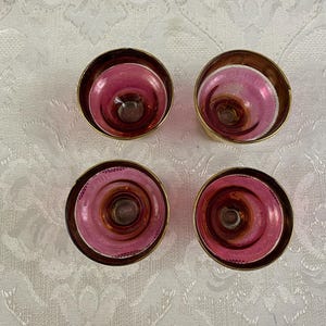 May include: Four vintage, pink and gold-rimmed glass candle holders. The candle holders have a circular design with a deep pink interior and a gold-colored rim. The candle holders are arranged on a white patterned surface.