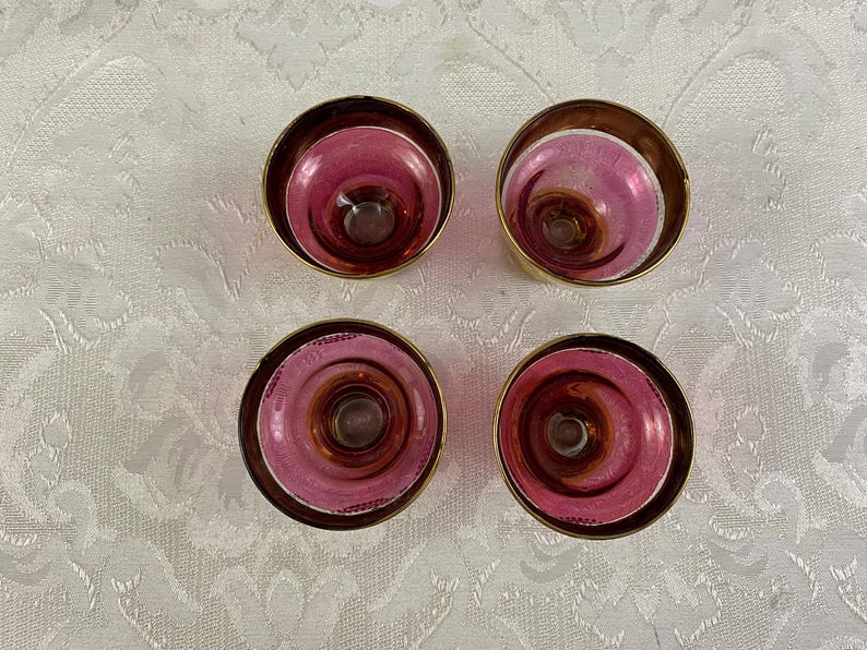 May include: Four vintage, pink and gold-rimmed glass candle holders. The candle holders have a circular design with a deep pink interior and a gold-colored rim. The candle holders are arranged on a white patterned surface.