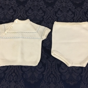 May include: A vintage white knit baby outfit, featuring a short-sleeved top with a textured yoke and button closure, paired with matching high-waisted shorts. The top has light blue accents. The outfit is displayed on a dark blue patterned surface.