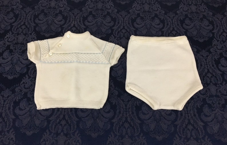 May include: A vintage white knit baby outfit, featuring a short-sleeved top with a textured yoke and button closure, paired with matching high-waisted shorts. The top has light blue accents. The outfit is displayed on a dark blue patterned surface.