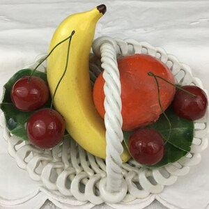 Może przedstawiać: Biały ceramiczny kosz na owoce z ż&oacute;łtym bananem, pomarańczową pomarańczą i czterema czerwonymi wiśniami.