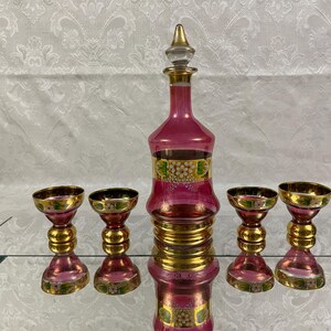 May include: A vintage pink glass decanter set with a stopper and four matching glasses. The set features gold accents and floral designs. The glasses and decanter have a pink hue with gold rims and bases.