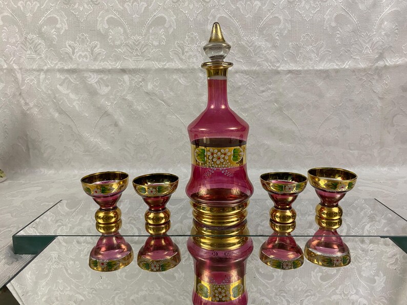 May include: A vintage pink glass decanter set with a stopper and four matching glasses. The set features gold accents and floral designs. The glasses and decanter have a pink hue with gold rims and bases.