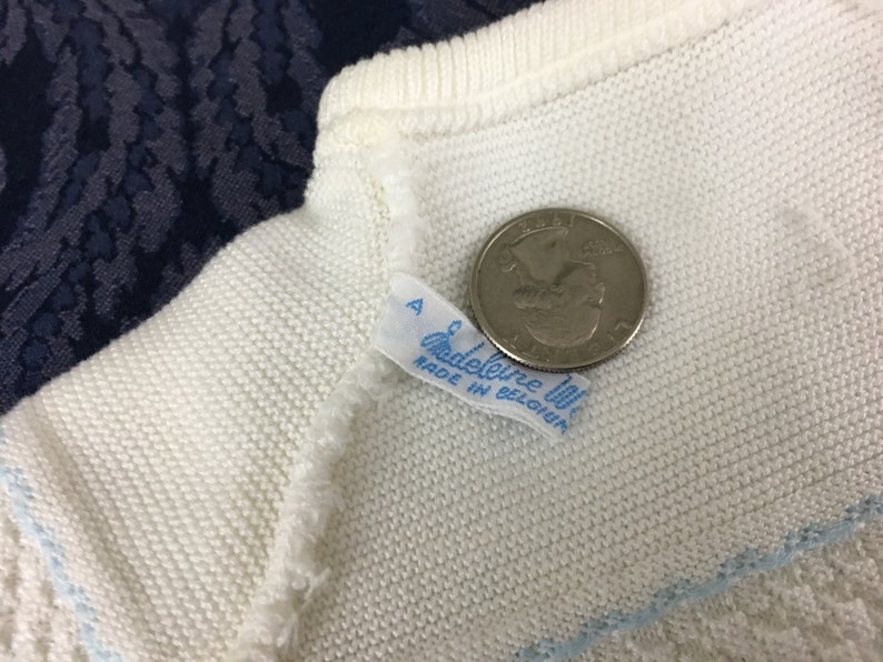 May include: Close-up of a white knitted garment with a light blue trim. A quarter coin is next to the garment for size comparison. A small tag reads "Madeleine We" and "Made in Belgium".