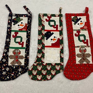 May include: Three Christmas stockings with a red, green, and black background. Each stocking features a snowman, gingerbread man, and the word "Joy" in a patchwork design.