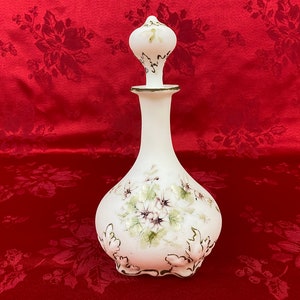 May include: White glass bottle with a floral design and a white stopper. The bottle has a bulbous body and a long, slender neck. The floral design is painted in shades of green and purple.