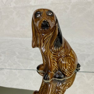 May include: A brown and black ceramic figurine of a dog sitting with its head tilted down. The dog has brown fur with black markings and brown eyes.