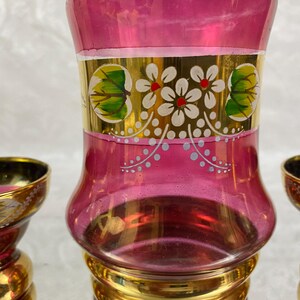 May include: A set of three decorative glass items. The central piece is a tall, pink vase with gold accents and floral designs. Two smaller matching pieces are on either side. The set has a vintage aesthetic.