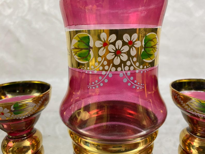 May include: A set of three decorative glass items. The central piece is a tall, pink vase with gold accents and floral designs. Two smaller matching pieces are on either side. The set has a vintage aesthetic.