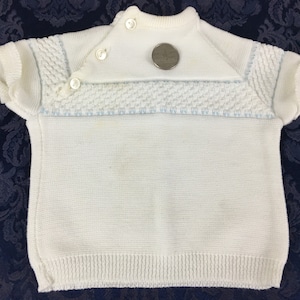May include: White knitted baby sweater with short sleeves. The sweater features a textured pattern across the chest and on the sleeves, with light blue accents. It has a button closure at the neckline and a ribbed hem. A quarter is placed on the sweater for size reference.