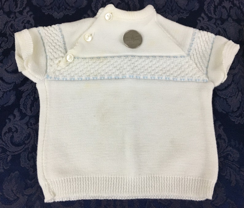 May include: White knitted baby sweater with short sleeves. The sweater features a textured pattern across the chest and on the sleeves, with light blue accents. It has a button closure at the neckline and a ribbed hem. A quarter is placed on the sweater for size reference.