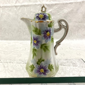May include: A porcelain coffee pot featuring a hand-painted floral design with purple and blue petals, yellow centers, and green leaves against a white background. The pot includes a matching lid with a finial, a curved handle, and gold trim.