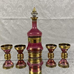 May include: A vintage decanter set featuring a ruby red glass decanter with a gold stopper and accents, accompanied by four matching goblets. The set is displayed on a reflective surface.